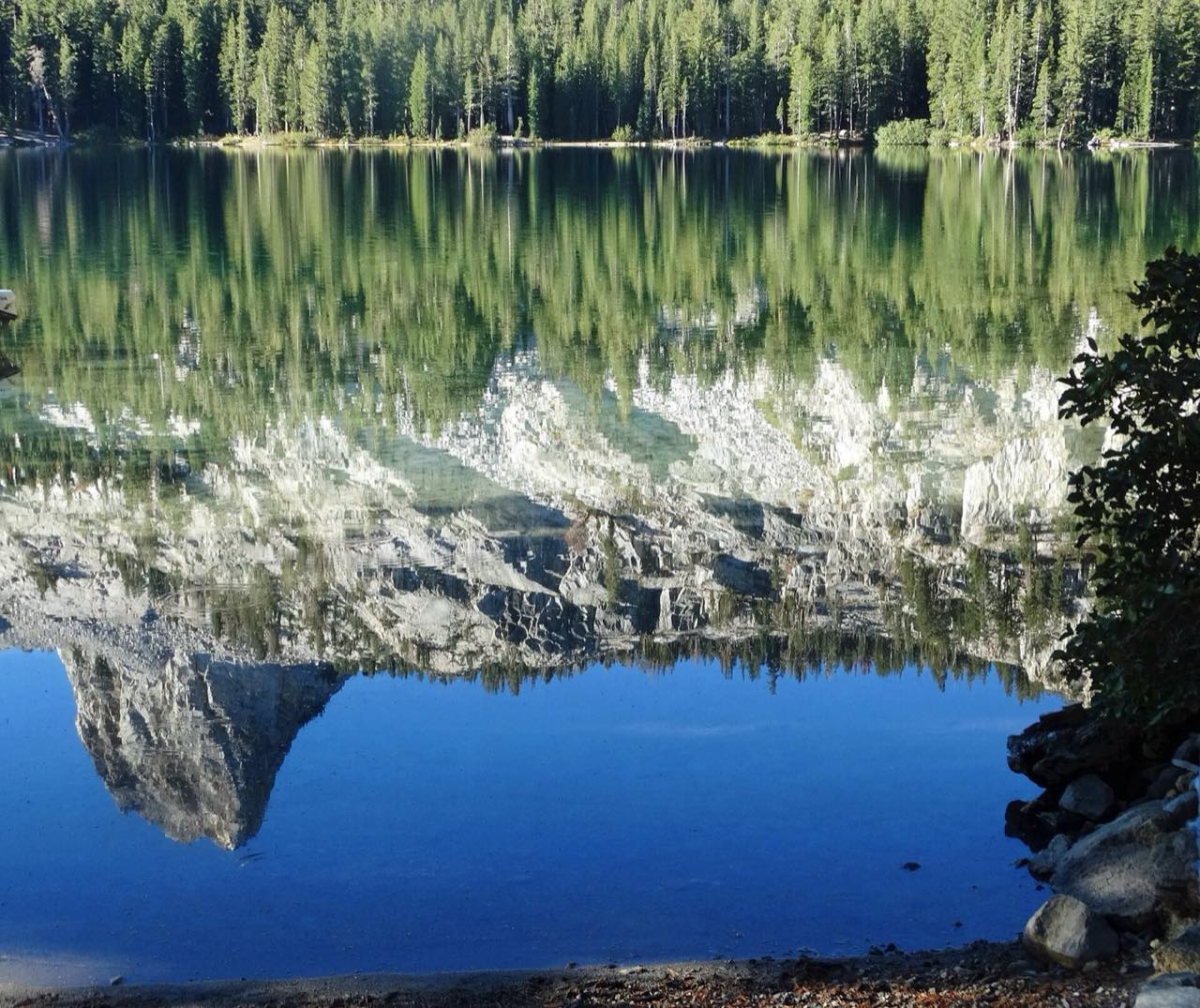 Lac de munte cu apă nemișcată care oglindește pădurea de brazi și versantul stâncos