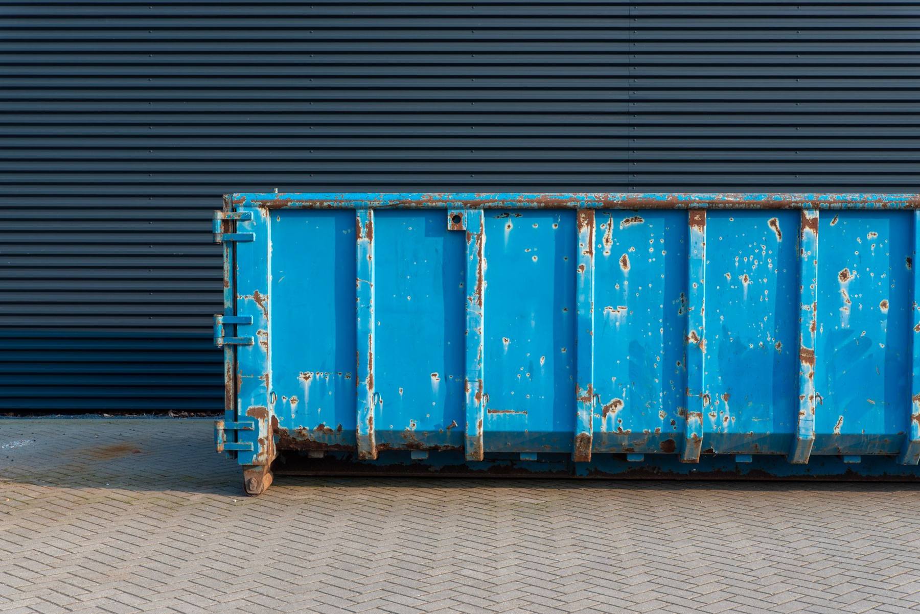 Un container abroll albastru, cu rugină pe margini, așezat pe pavaj în fața unui perete de tablă cutată