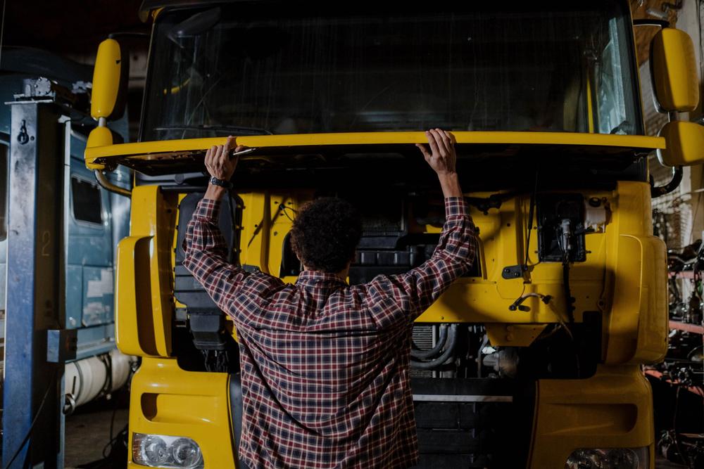 Un om în cămașă în carouri ține deasupra capului capota deschisă a unui camion galben; dedesubt se văd radiatorul și furtunurile.