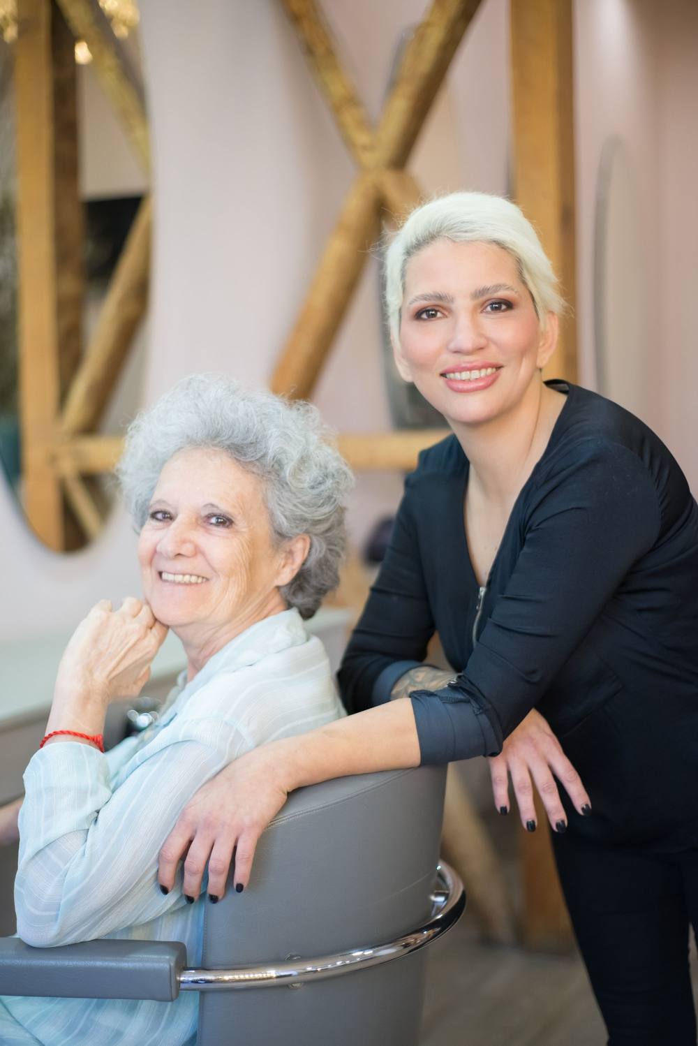 Doamna Lia, cu părul cărunt creț, stă în scaun și zâmbește; lângă ea, Sorina, cu părul platinat scurt, sprijinită de spătar. În spate, grinzile de lemn în X.