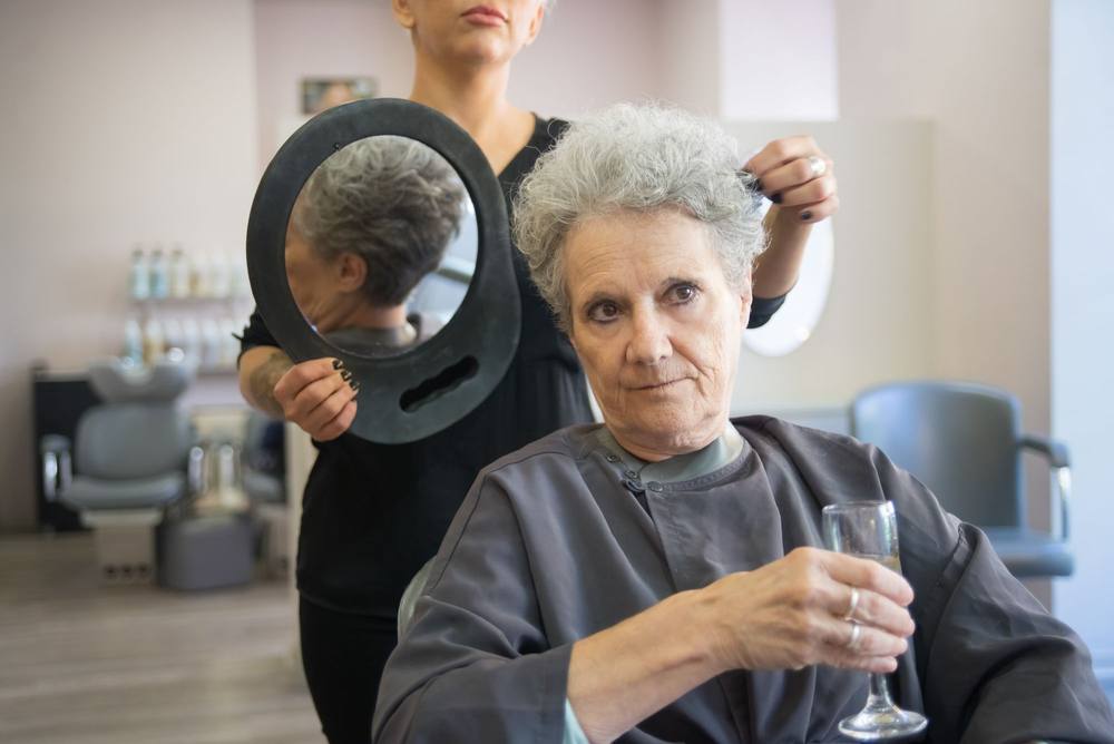 Doamna Lia, cu un pahar în mână, se uită în oglindă; Sorina ține în spatele capului ei o oglindă rotundă de mână.