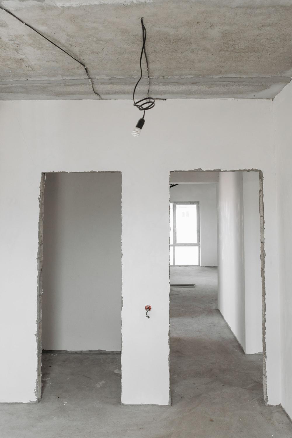 Două goluri de ușă cu marginile de zidărie neterminate, un bec atârnat de un cablu din tavanul de beton și șapa goală pe jos.