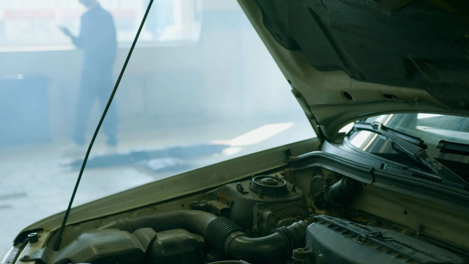 Compartimentul motorului unei mașini cu capota ridicată; în spate, în hala luminată, un om neclar se uită la telefon.