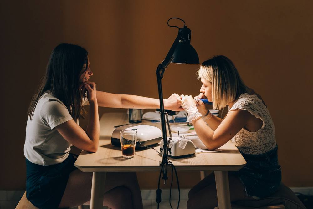 Seara, la masa de lemn: clienta brunetă își sprijină bărbia în palmă și întinde mâna; Irina, cu mănuși, îi lucrează unghiile sub lampa neagră.
