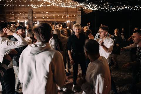 Invitați care dansează sub becuri, în fața unei barăci de lemn