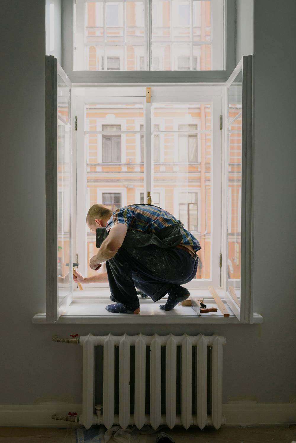Un tâmplar ghemuit pe pervazul unei ferestre înalte, cu cercevelele deschise; afară, fațada galbenă a casei de vizavi.
