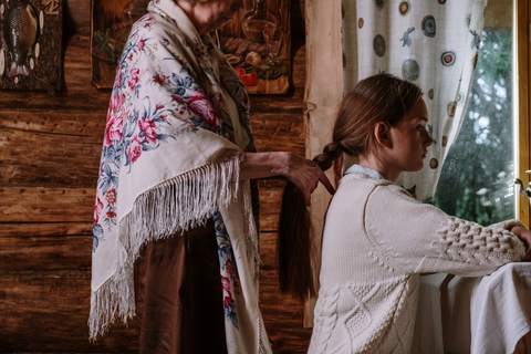 Gazda, cu un șal cu trandafiri pe umeri, împletește părul lung al unei fete care stă la fereastră