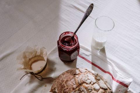 Pe o față de masă albă cu dungă roșie: un borcan de dulceață cu lingura în el, un pahar cu lapte și o pâine rotundă