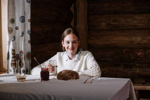 O fată în pulover alb stă singură la o masă lungă cu față de masă albă, cu un pahar de lapte, un borcan de dulceață și o pâine