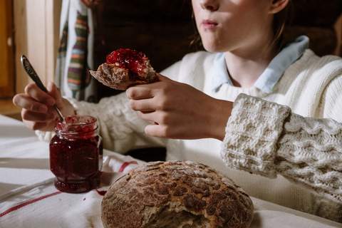 O fată unge cu dulceață de zmeură o bucată de pâine, cu borcanul și pâinea rotundă pe masă