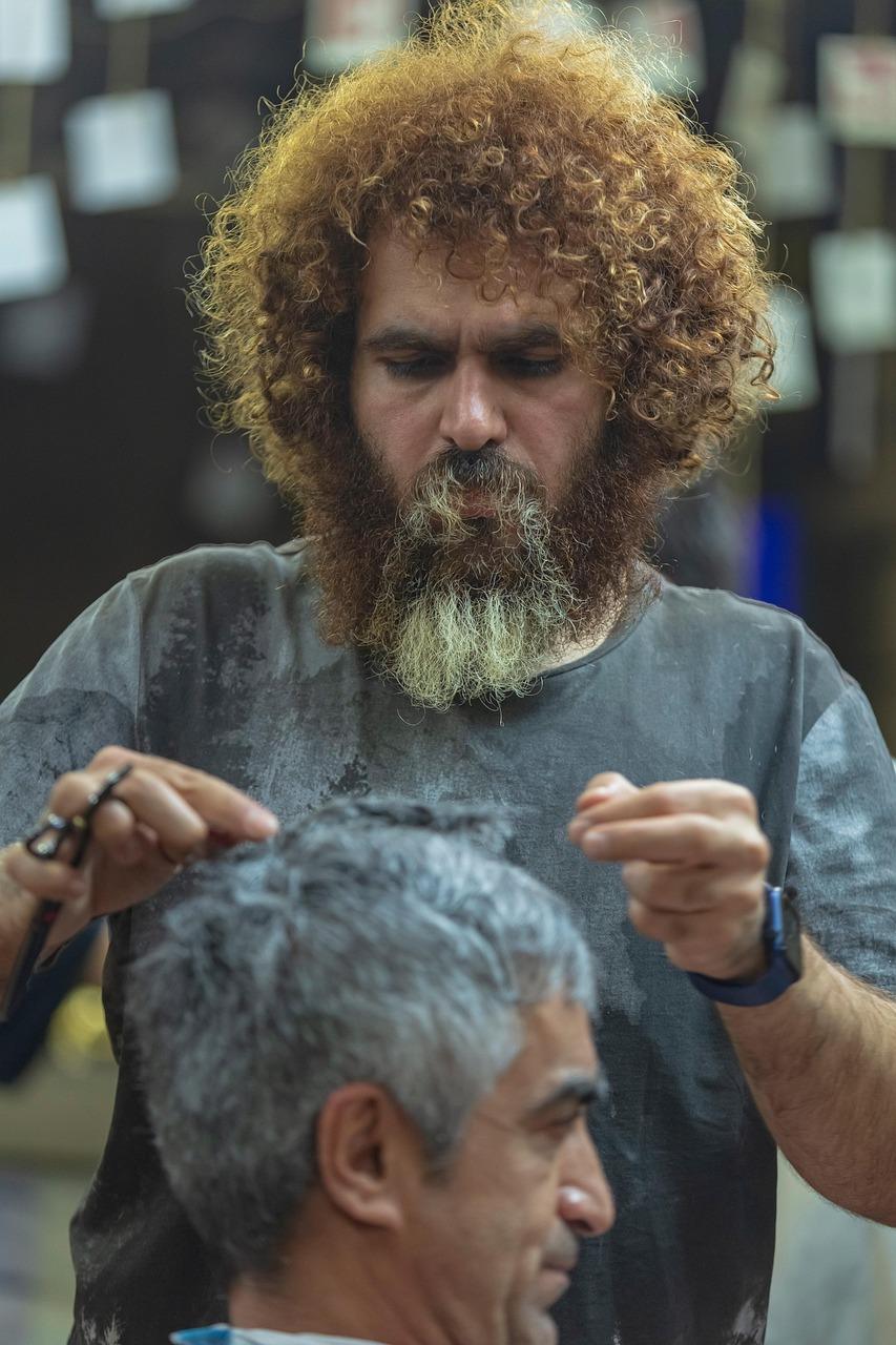 Arman, cu păr roșcat creț și barba căruntă spre vârf, tunde cu foarfeca un domn cu păr alb.
