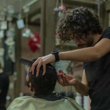 Darius ține pieptenele pe creștetul unui client și foarfeca în cealaltă mână; în spate, poze pe sfori.