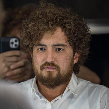 Un tânăr cu păr creț și cămașă albă privește în oglindă; cineva ține un telefon lângă el.