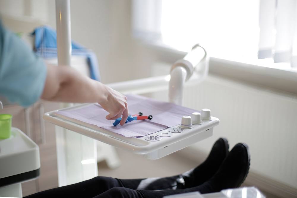Tava cu instrumente acoperită cu hârtie lila; o mână în mănușă alege o seringă, iar în colț se văd picioarele pacientei pe scaun.