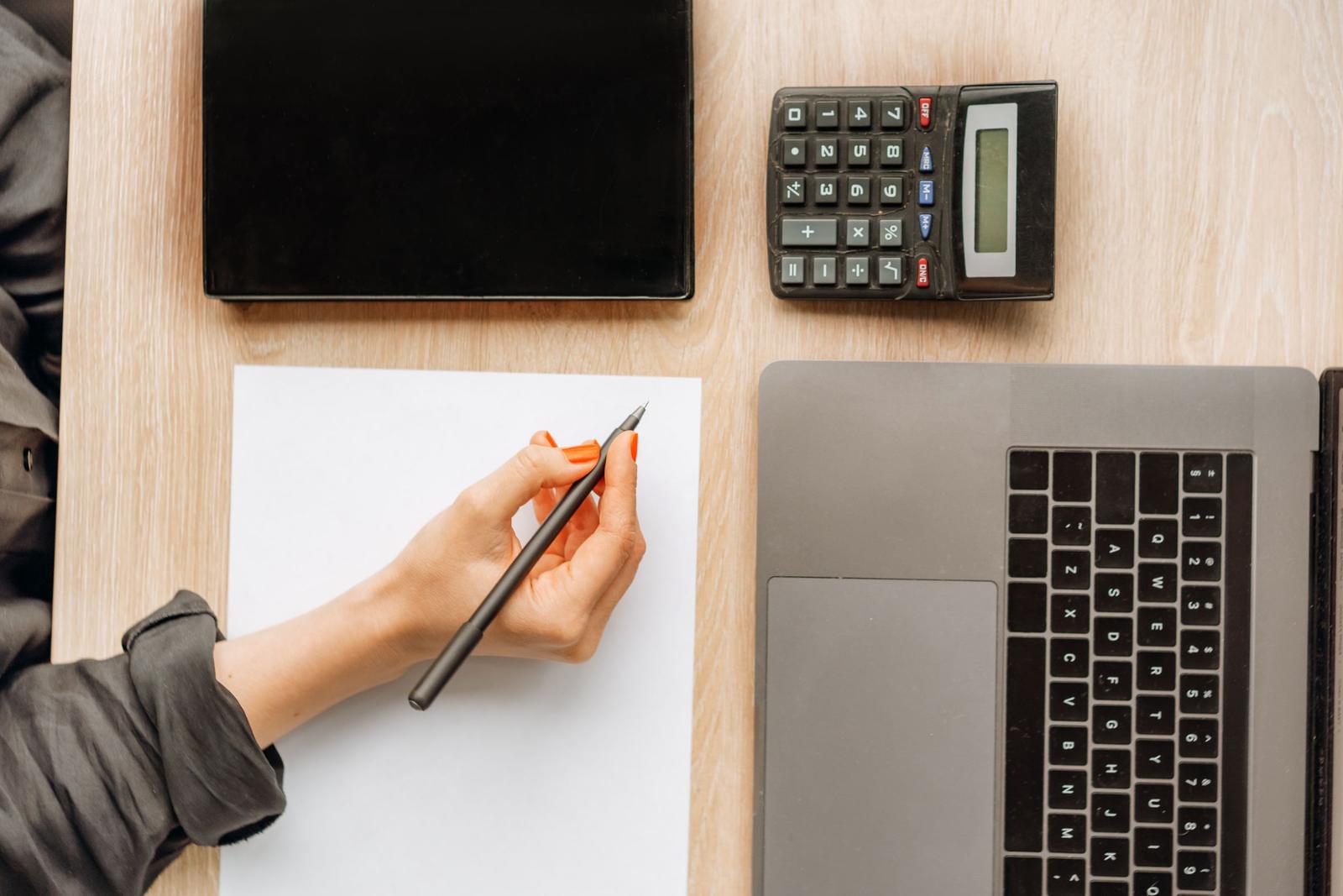 Birou văzut de sus: o mână cu pixul deasupra unei foi albe, lângă un calculator, o agendă neagră și un laptop.