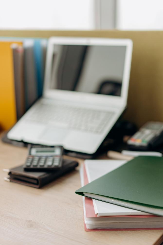 Colț de birou: un dosar verde peste teancuri de foi, un calculator pe o agendă, laptopul și bibliorafturile lângă fereastră.